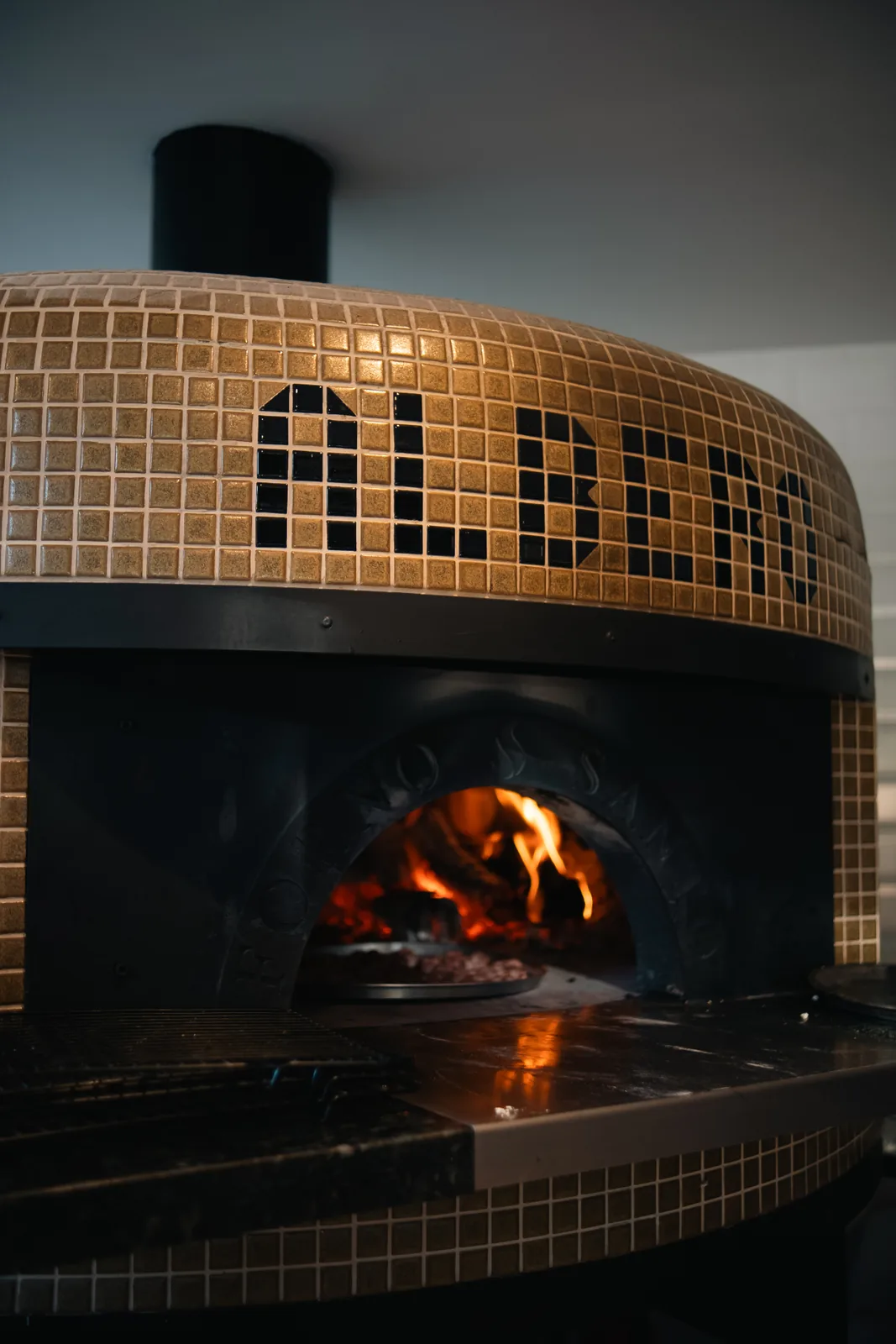 Forno à lenha revestido em mosaico dourado com o nome ALBERO em pastilhas escuras.